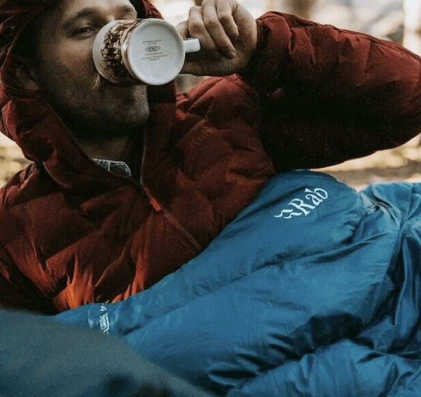 man lying down drinking coffee from mug in rab sleeping bag