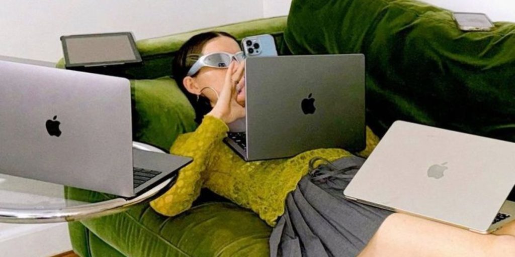girl in green laying on green sofa on phone surrounded by Apple laptops and a tablet