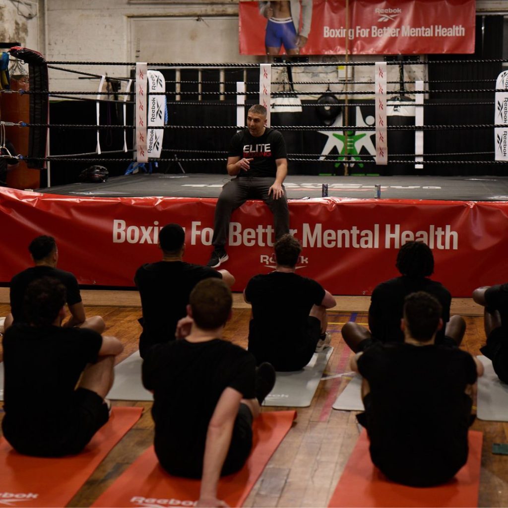 A group of men sit on yoga mats in front of a boxing ring listening to an Elite Community Hub coach speak at a Reebok boxing event.