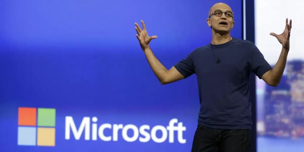 Satya Nadella speaking at a conference with the Microsoft logo on a screen in the background.